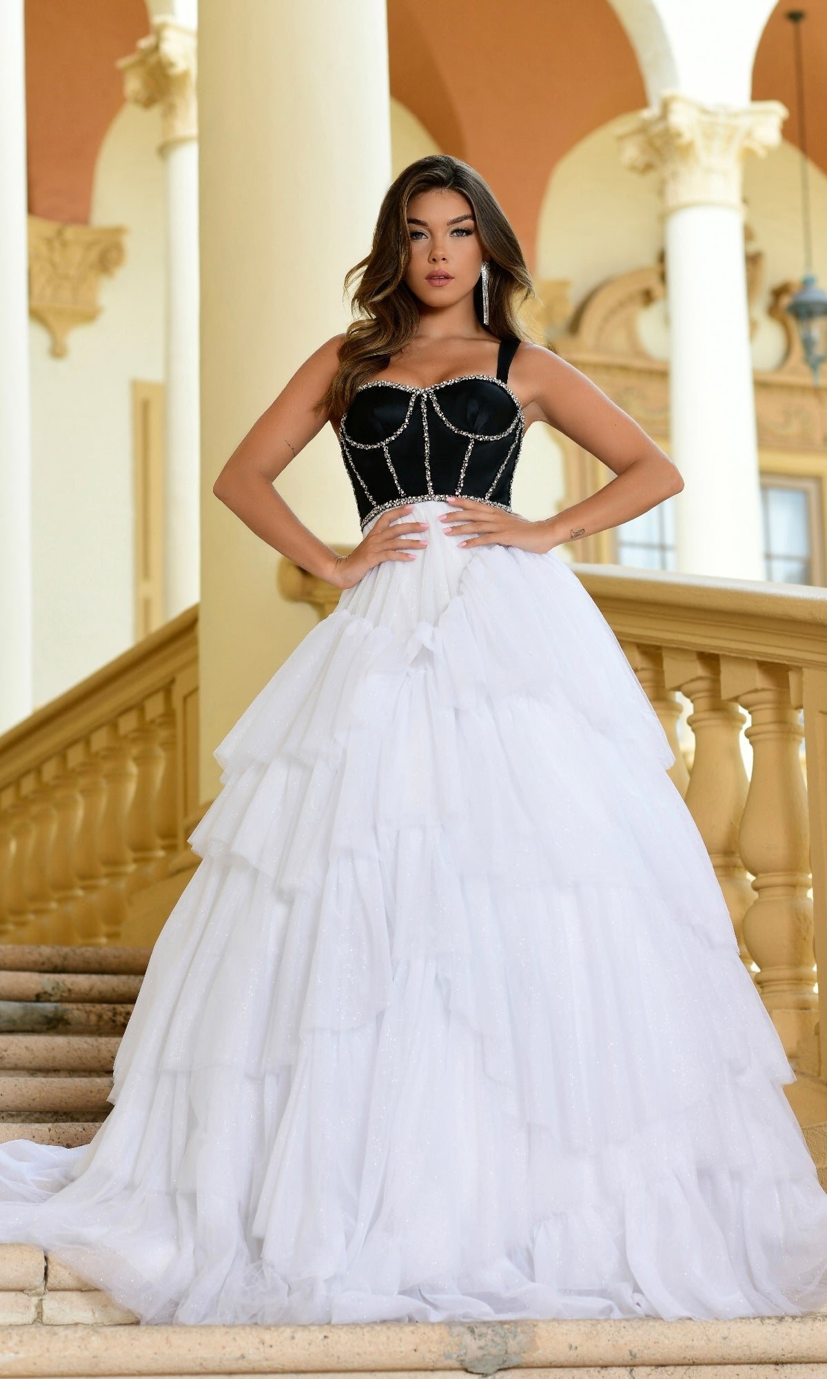 A woman stands on a staircase wearing the Long Formal Dress 28592 by Ava Presley, featuring a strapless black corset bodice and a voluminous white tulle A-line skirt.